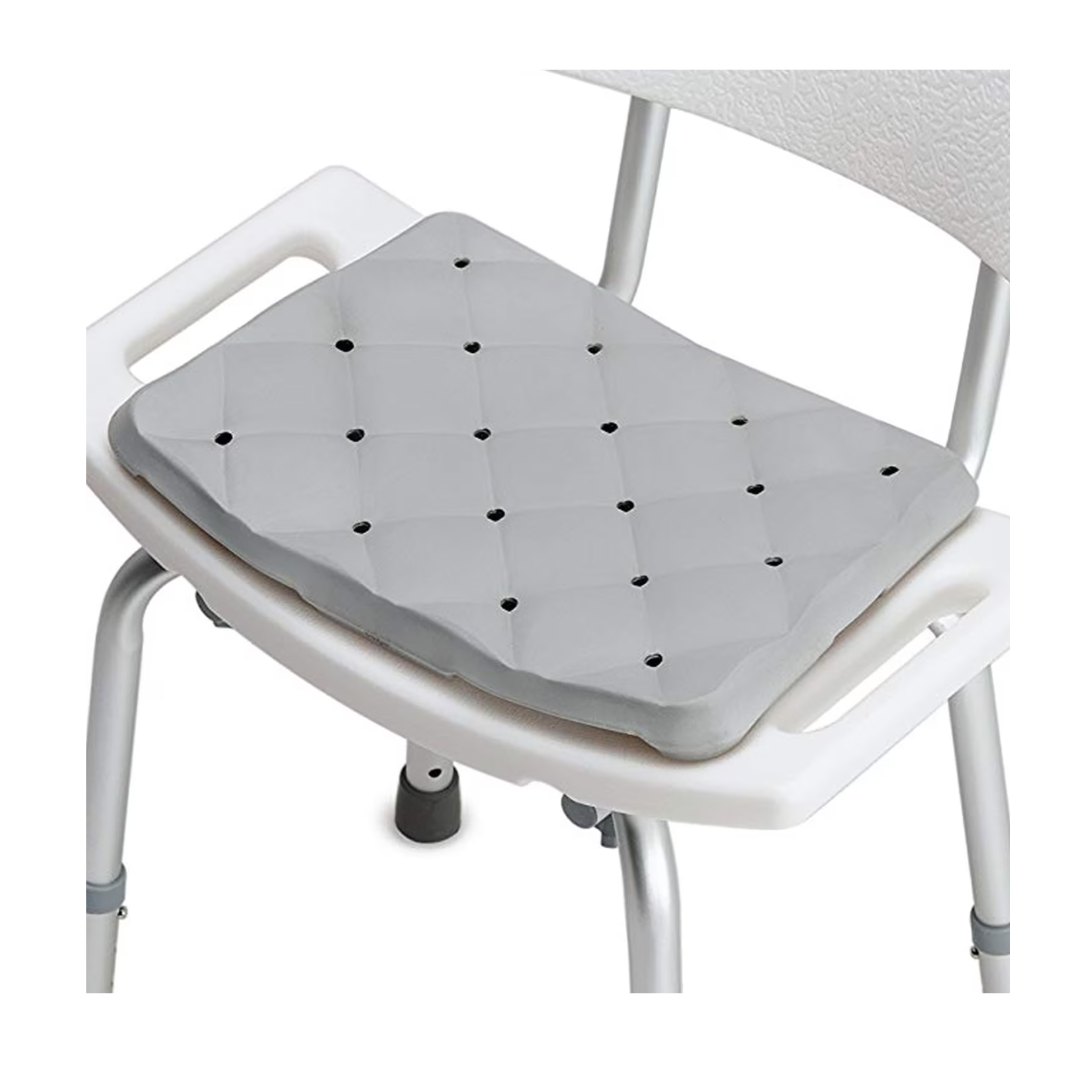 The DMI Waterproof Foam Bath Seat Cushion, gray and quilted with small ventilation holes for slip prevention, is shown on a white shower chair with armrests and metal legs.